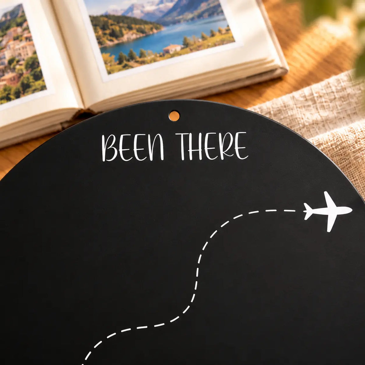 Illustrative product detail: Close-up of the black Circle Travel Board showing white Been There lettering, the dashed flight route, airplane motif and empty top mounting hole.